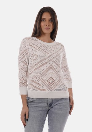 Donna con capelli lunghi e scuri che indossa un maglione bianco lavorato a maglia con motivo e jeans grigio chiaro, in piedi davanti a uno sfondo semplice.