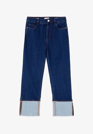 Siniset farkut leveillä käänteillä, jotka paljastavat vaaleamman sisäpuolen kankaan ja sivuvetoketjut käänteissä, etutaskuissa ja napinauhassa.