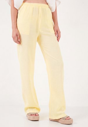 Lockere hellgelbe Hose mit elastischem Bund und Kordelzug, getragen mit beigen Plateausandalen und einem weißen Top, das am Taillenbund sichtbar ist.