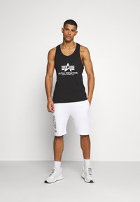 Schwarzes Tanktop mit weißem "Alpha Industries" Logo, kombiniert mit weißen Shorts mit goldenem Markendruck. Model trägt weiße Sneaker.