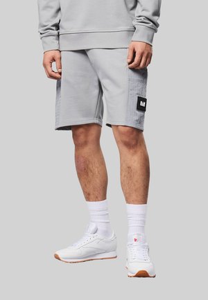 Mężczyzna w jasnoszarych szortach z boczną kieszenią, białych skarpetkach i białych tenisówkach na jednolitym szarym tle.