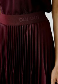 Burgunderfarbene, gefaltete Bluse mit einem breiten, mit dem Text "OUI OUI" versehenen elastischen Bund. Glattes, glänzendes Material mit weicher Textur.