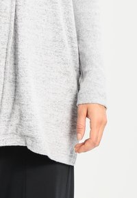 Grå stickad tröja med långa ärmar, en lös passform och rundad nederkant. Tygets textur verkar mjuk och lite melerad.