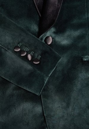 Blazer de terciopelo verde oscuro con cuello de chal de satén, acentos de botones en los puños y una textura suave con un ligero brillo.