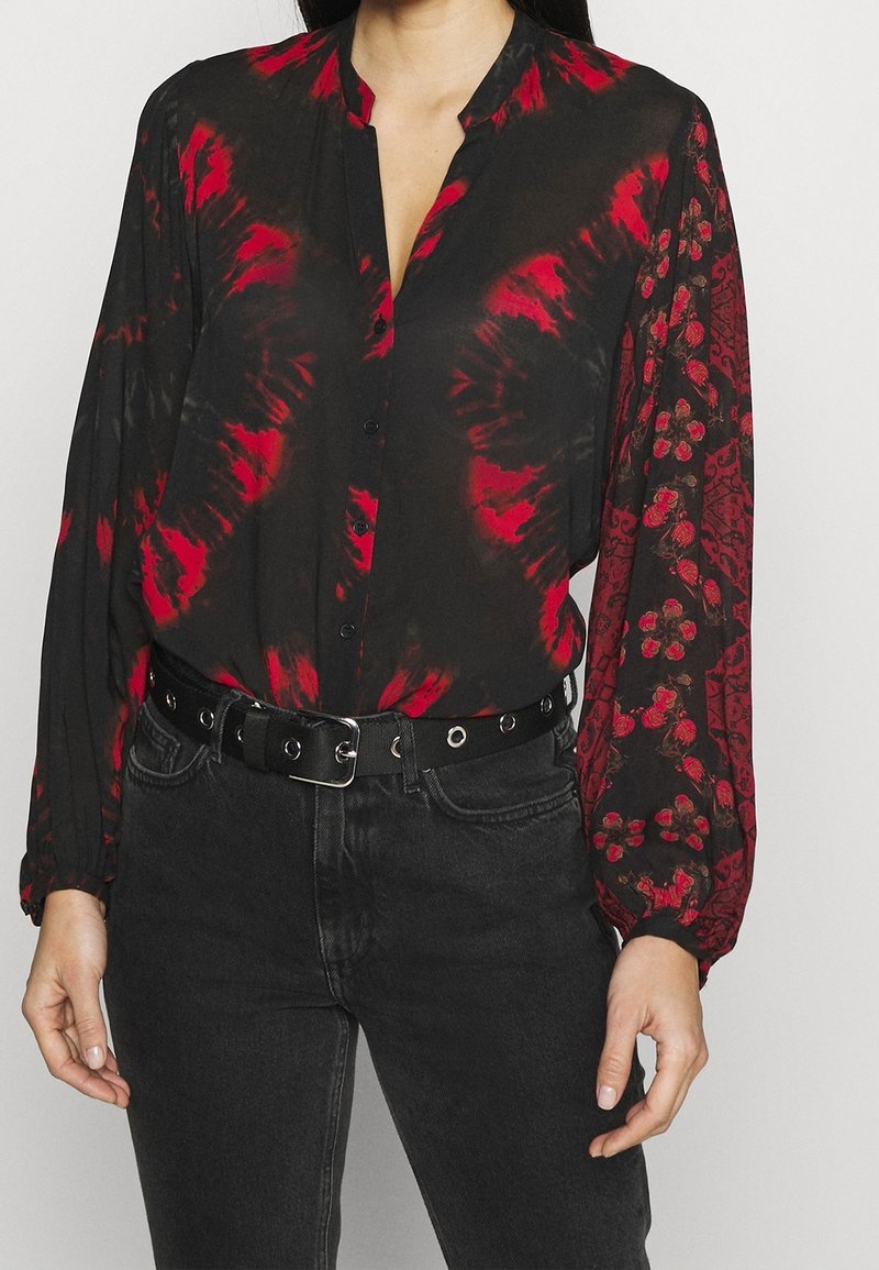 Blouse noire à boutons avec motif tie-dye rouge et dessins floraux sur les manches, associée à un jean noir et une ceinture noire à œillets argentés.