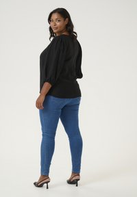 Bluză neagră texturată cu mâneci bufante, asociată cu blugi skinny albaștri și sandale negre cu toc înalt. Fundal simplu și curat.