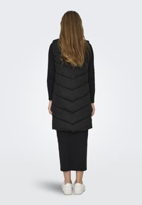 Gilet matelassé noir avec un motif en chevrons, s'étendant jusqu'au milieu de la cuisse, porté sur un haut à manches longues noir et une jupe, associé à des baskets blanches.