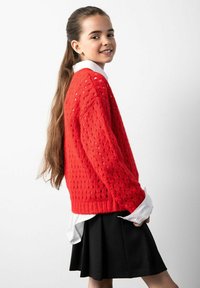 Czerwony sweter z dzianiny o luźnym splocie, zestawiony z białą koszulą z kołnierzykiem pod spodem oraz czarną spódnicą. Długie włosy spływają na plecy.