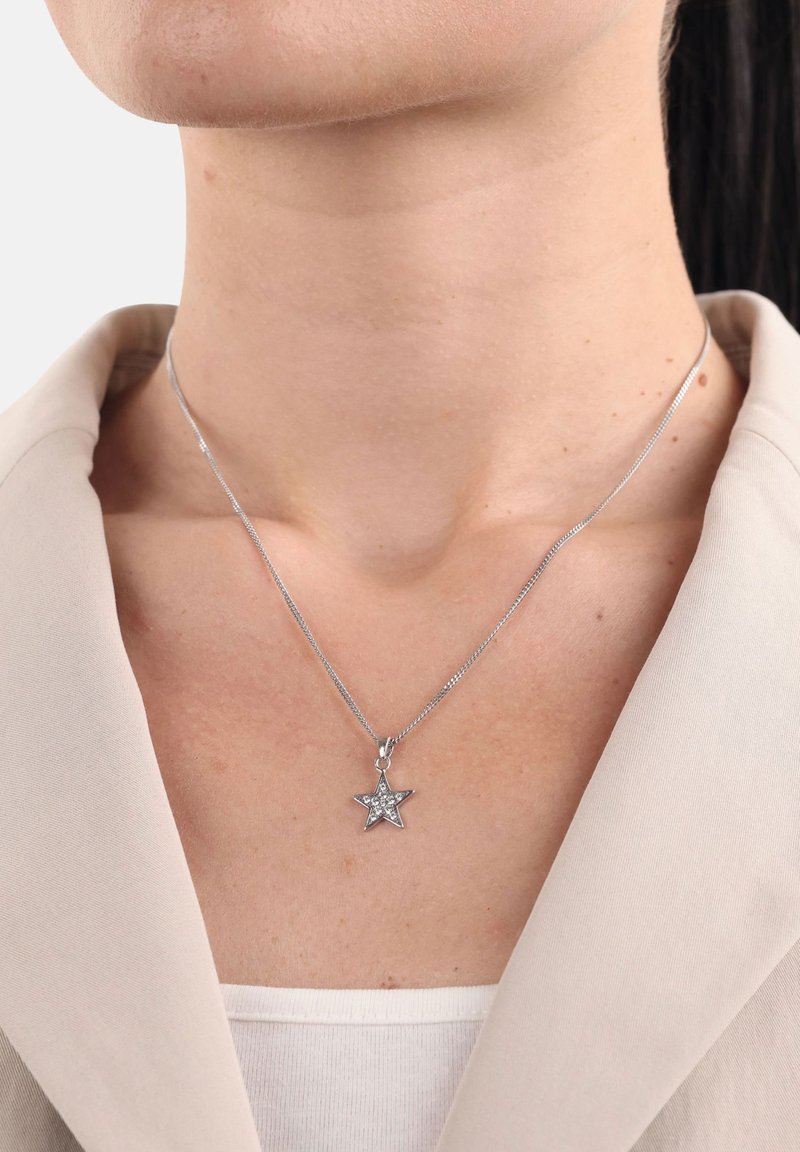 Silvrig kedja med en stjärnanhäng som pryds av klara rhinestones, bärs över en ljusfärgad topp, mot en neutral bakgrund.