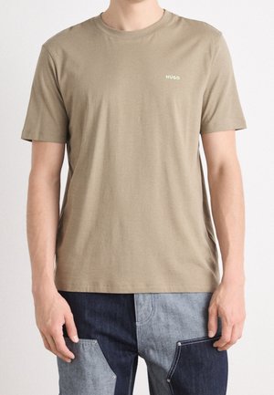 Mand iført plain beige T-shirt med korte ærmer og lille "HUGO" logo på brystet, kombineret med tofarvede patchwork denim jeans.