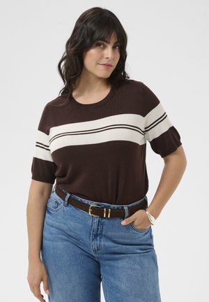 Femme aux cheveux foncés portant un pull marron à manches courtes avec des rayures blanches et un jean bleu, debout, une main dans la poche.