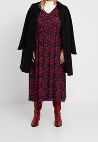 Vestido azul marino con patrón floral rojo, hasta la rodilla, con cintura ajustada, combinado con un abrigo negro y botas rojas hasta la rodilla. Jersey de cuello alto blanco debajo.