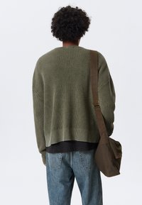 Olive green knitted sweater with a textured pattern, cropped hem, and dropped shoulders, paired with blue denim jeans and a brown messenger bag.