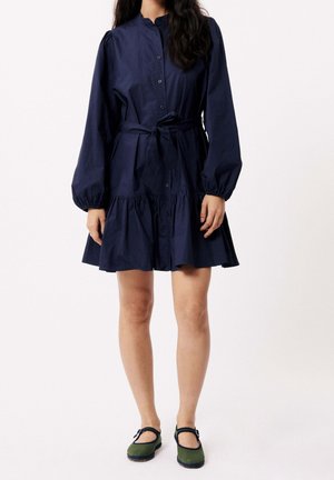 Femme portant une robe bleu marine à manches longues, boutonnée sur le devant, avec une ceinture à la taille, et des chaussures Mary Jane vertes, debout devant un fond blanc.