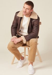 Braune Lederjacke mit(beigem) Schafpelzkragen, getragen über einem weißen Strickpullover, kombiniert mit beigefarbenen Cordhosen und weißen Sneakers, sitzend auf einem Hocker.