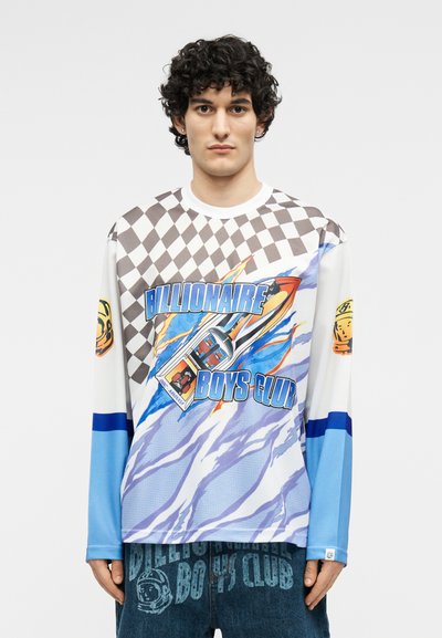 Νεαρός άνδρας με σγουρά μαλλιά φοράει μια πολύχρωμη μπλούζα μακρυμάνικη Billionaire Boys Club με μοτίβο σκακιέρας και γραφικό σχέδιο ρουκέτας.