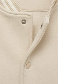 Nahaufnahme von beigem Stoff, der einen teilweise geöffneten Druckknopfverschluss zeigt, mit einem Metall- und einem beigen Knopf an einem gestrickten Kleidungsstück.