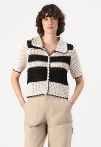 Schwarz und cremefarbene gehäkelte Kurzarm-Cardigan mit gewelltem Kragen und Knopfverschluss, der gestreifte Muster und ein offenes Strickdesign aufweist.