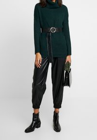 Donkergroen geribbelde gebreide trui met een col, gecombineerd met zwarte glanzende taps toelopende broek. Accessoires omvatten een zwarte riem en handtas.