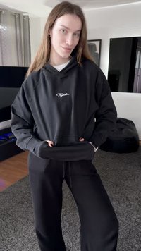 Melns džemperis ar kapuci, uz tā ir logotips, papildināts ar ribām uztvertām aprocēm un priekšējo kabatu, kopā ar melnām sporta biksēm. Audums šķiet mīksts un ērts.