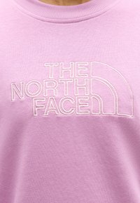 Detailná záber svetlofialovej mikiny s vyšitou bielou niťou nápisom "THE NORTH FACE" na oblasti hrude.