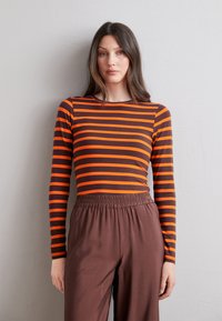 Dlhý rukáv top s horizontálnymi prúžkami v oranžovej a tmavohnedej farbe, v kombinácii s hnedými nohavicami s elastickým pásom. Hladká textúra látky.