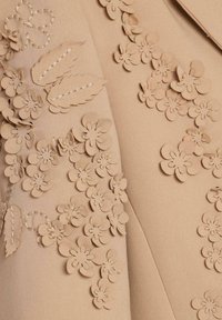 Beiger Stoff mit 3D-Blumenapplikationen in verschiedenen Größen, mit gesteppten Details und glatter Textur. Fokus auf florale Verzierungen.