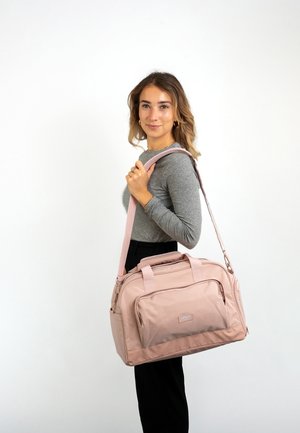 Rosa stof duffelbag med dobbelthåndtag og skulderrem, der har en frontlomme og en glat tekstur. Modellen holder tasken på skulderen.
