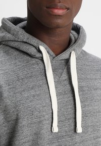 Sudadera gris con una textura suave, que cuenta con un cierre de cordón en blanco roto y un escote en V, destacando un diseño casual.