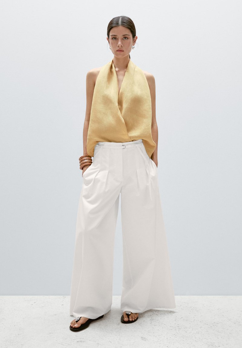 Satintopp i gul med draperad design, kombinerad med vita vida byxor med hög midja. Modellen bär bruna slip-in-sandaler och träarmband.