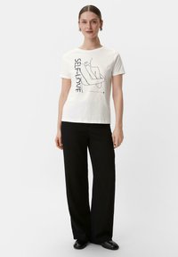 Weißes Baumwoll-T-Shirt mit einer schwarzen Strichzeichnung und dem Text "SELBSTLIEBE." Kombiniert mit weitgeschnittenen schwarzen Hosen und schwarzen Schuhen. Einfaches, lässiges Design.