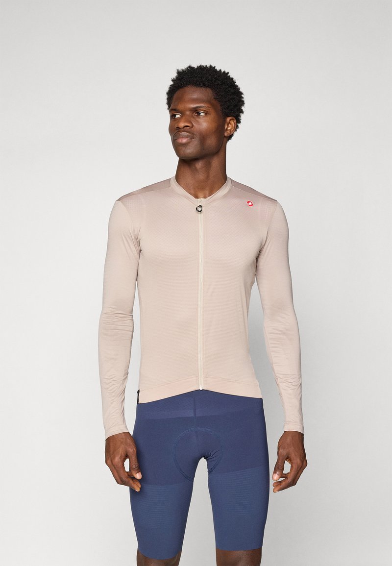 Beige langärmliges Radtrikot mit Reißverschluss vorne, ausgestattet mit einem strukturierten Design und kleinem Logo. Getragen mit blauen gepolsterten Radhosen.