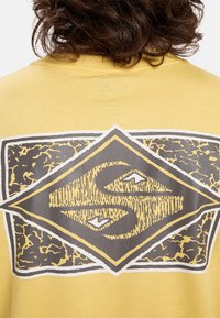 Žluté tričko s velkým grafickým prvkem na zádech ve tvaru diamantu. Design obsahuje texturované šedé a černé prvky s bílými konturami.