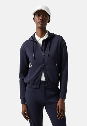 Kobieta w granatowej bluzie z kapturem na zamek błyskawiczny i dopasowanych spodniach, z białą koszulą kołnierzykową oraz białą czapką bejsbolową, stojąca na tle jednolitego tła.