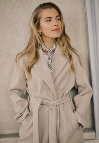 Bež plašč iz mešanice volne z pasom v pasu, širokimi ovratniki in stranskimi žepi; dopolnjen s črno-belim črtastim šalom.