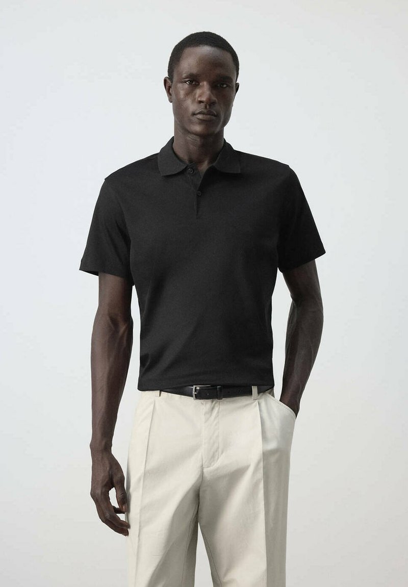 Homme portant un polo noir à manches courtes rentré dans un pantalon beige clair avec une ceinture noire, debout devant un fond gris uni.