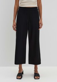 Pantalon noir avec une coupe évasée, fabriqué en tissu lisse. Caractérisé par une taille haute et une coupe décontractée, associé à des sandales à glissière noires.
