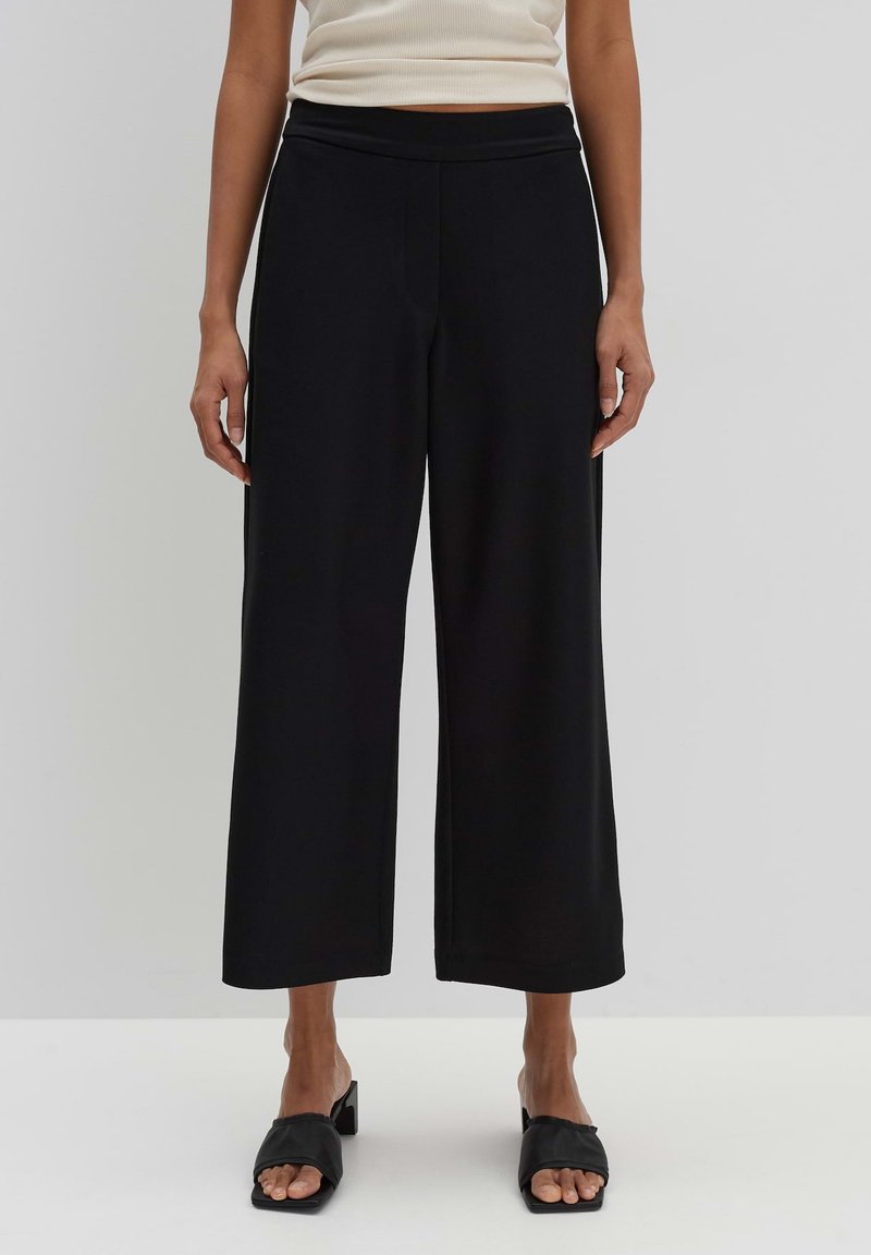 Pantalon noir avec une coupe évasée, fabriqué en tissu lisse. Caractérisé par une taille haute et une coupe décontractée, associé à des sandales à glissière noires.