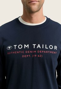 Camisa de manga larga de color navy hecha de algodón. Cuenta con un cuello redondo blanco y un estampado en la parte frontal: "TOM TAILOR AUTHENTIC DENIM DEPARTMENT" en letras gruesas.