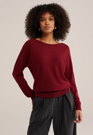 Donna con capelli ricci che indossa un top bordeaux a maniche lunghe e pantaloni a vita alta grigio scuro a righe sottili, in piedi davanti a uno sfondo neutro.