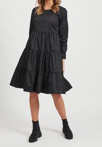 Schwarzes, langärmeliges Kleid mit einem gestuften Design, gefertigt aus einem glatten Stoff. Verfügt über eine geraffte Taille und einen gerafften Saum, kombiniert mit schwarzen Stiefeln bis zum Knöchel.