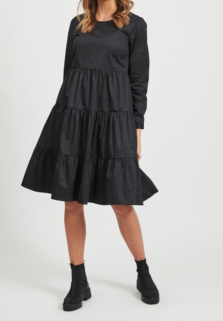 Schwarzes, langärmeliges Kleid mit einem gestuften Design, gefertigt aus einem glatten Stoff. Verfügt über eine geraffte Taille und einen gerafften Saum, kombiniert mit schwarzen Stiefeln bis zum Knöchel.