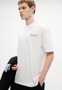 Bílý bavlněný tričko s černým logem "Hugo" na levé hrudi. Model nosí černou bundu, která ukazuje uvolněný střih a kulatý výstřih.