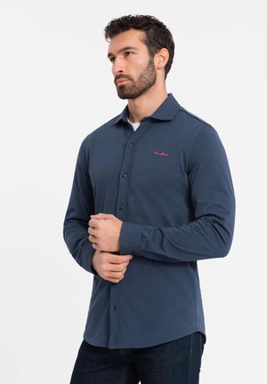 Marineblaue Langarmbluse mit Kragen, die Knöpfe vorne und ein gesticktes rotes Logo auf der linken Brust hat. Weiche Stofftextur.