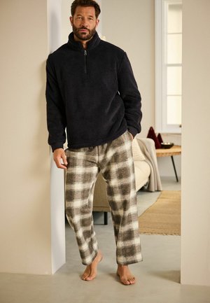 Mustat fleece-takit, joissa on puoli vetoketju, yhdistetty vaaleanruskeisiin ja kermaisiin ruutukuosisisiin pyjamasin housuihin. Pehmeät kankaat ja rento istuvuus. Paljain jaloin neutraalissa ympäristössä.