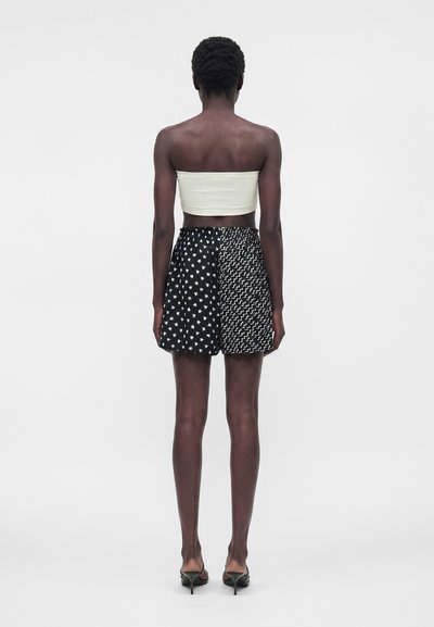 Γυναίκα που στέκεται με την πλάτη, φοράει λευκό crop top χωρίς τιράντες, μαύρα σορτς με δύο διαφορετικά σχέδια και μαύρα πέδιλα με τακούνι.