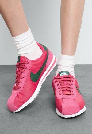 Rózsaszín Nike sportcipők zöld kiegészítőkkel, sima és velúr textúrák keverékével, kerek orral és fehér gumitalppal, amelynek fogazott szélei vannak.