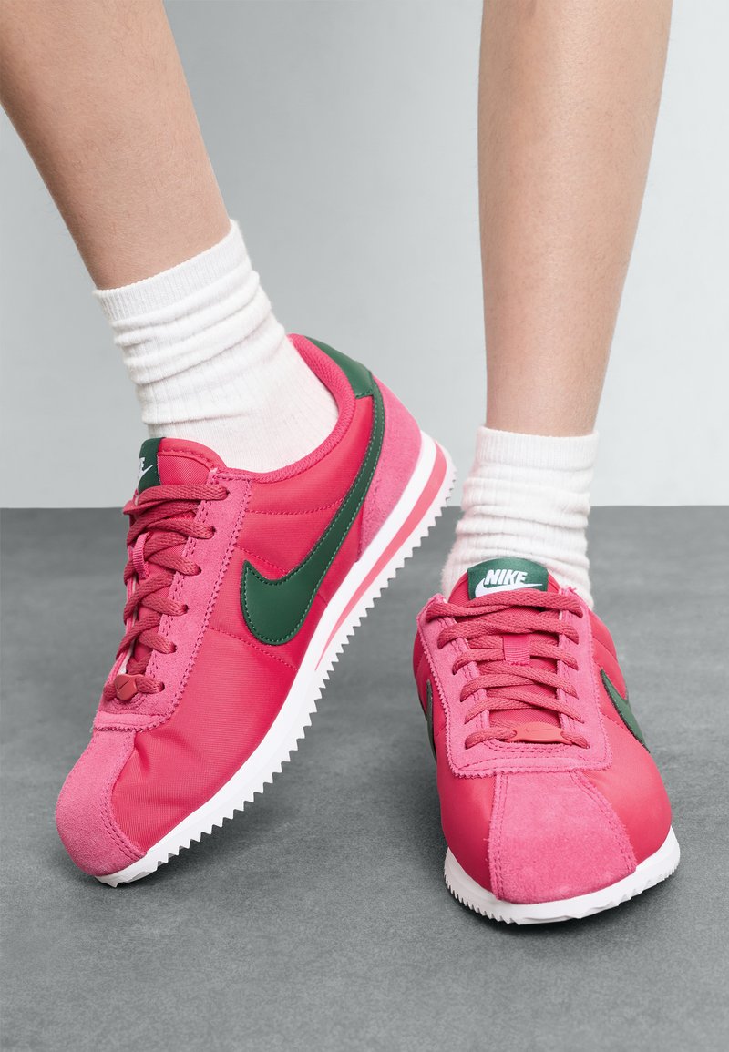 Ružičaste Nike tenisice s zelenim akcentima, koje se ističu kombinacijom glatke i seme teksture, okruglog vrha te bijelim gumenim potplatom sa nazubljenim rubovima.