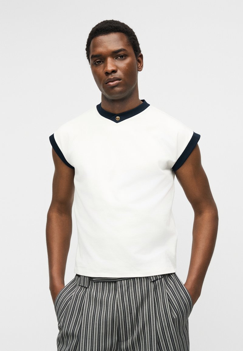 Man wearing sleeveless white shirt with black trim and gold button, paired with gray and black striped pants, hands in pockets.