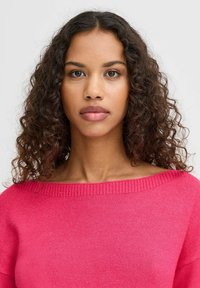 Pull en maille d'un rose vif avec un large col et des poignets côtelés, présentant une texture douce et une coupe décontractée, porté par un modèle aux cheveux bouclés.